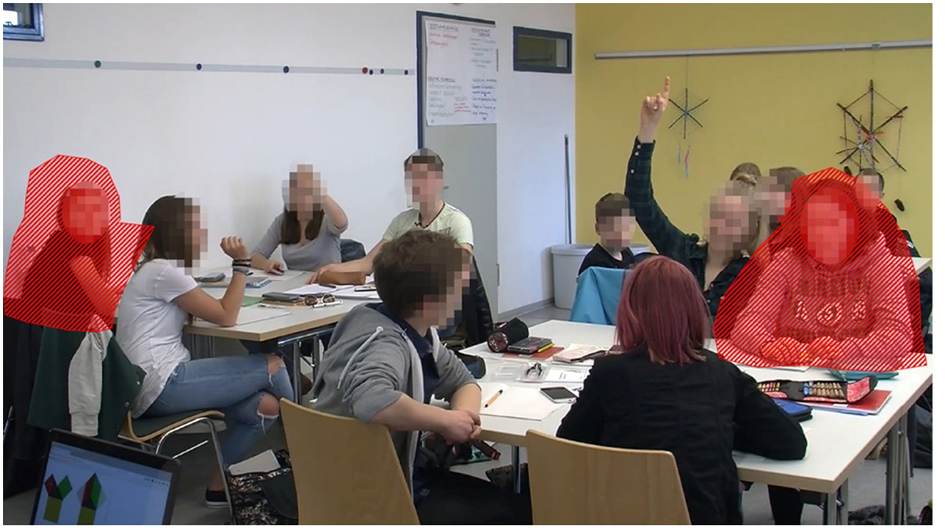 A group of students sits around a table in a classroom. Two students outlined in red, one on the left and one on the right, are highlighted. Another student raises a hand, signaling a question or contribution. The students' faces are pixelated for privacy. The classroom has white and yellow walls with educational materials displayed.