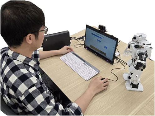 Person wearing glasses and a checkered shirt sits at a desk using a computer. A small humanoid robot stands beside the monitor, which displays a webpage.