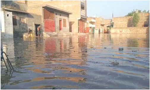 Flooded street with murky water reflecting nearby buildings and houses. A few people stand near a doorway, with scattered debris floating on the surface.