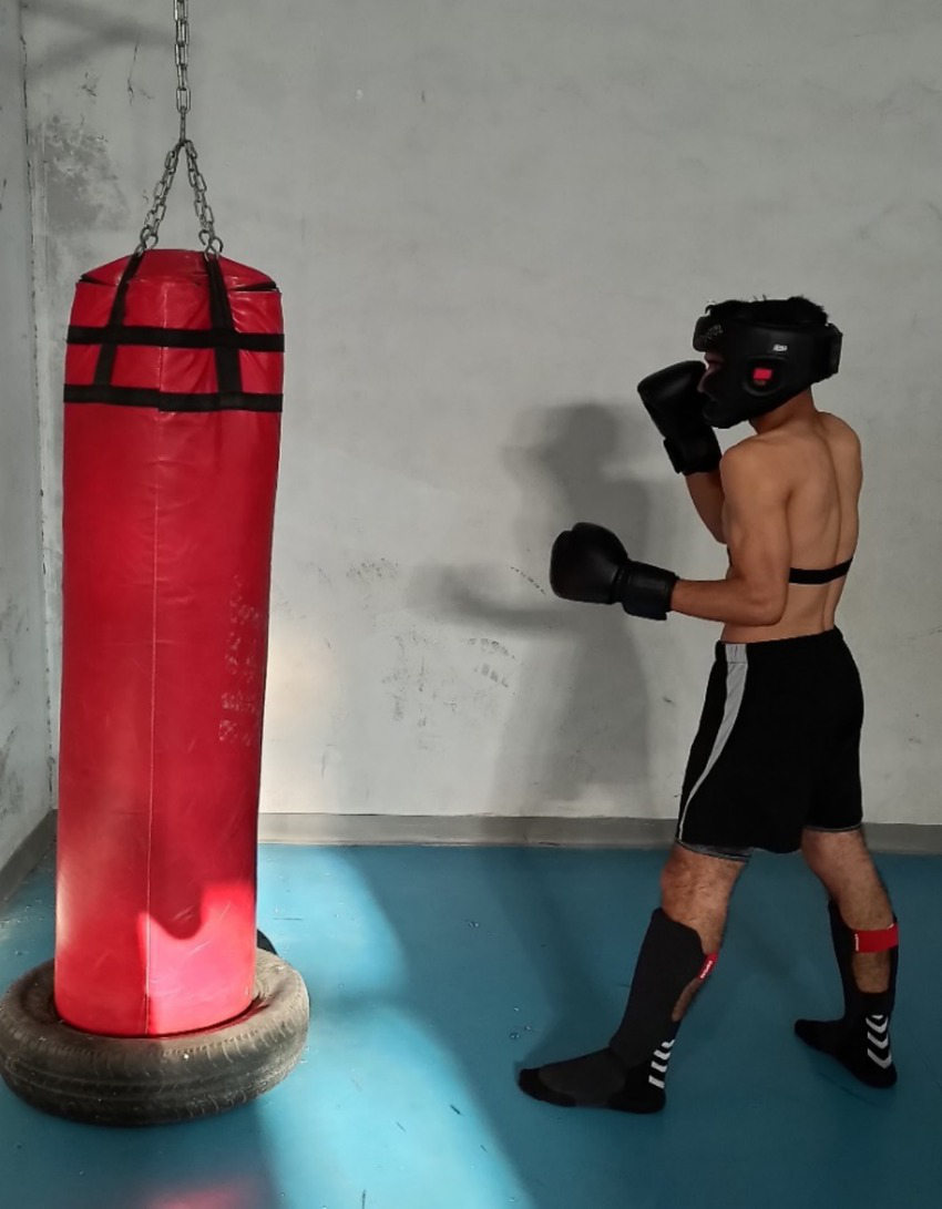 A person in boxing gear, including gloves and a protective helmet, stands facing a suspended red punching bag. The setting is a room with a minimalist, industrial style, featuring a concrete wall and a blue floor.