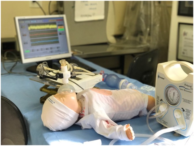 A medical training setup with a baby mannequin on a table, connected to a breathing apparatus and monitor. The monitor displays vital signs, and a resuscitator device is visible nearby.