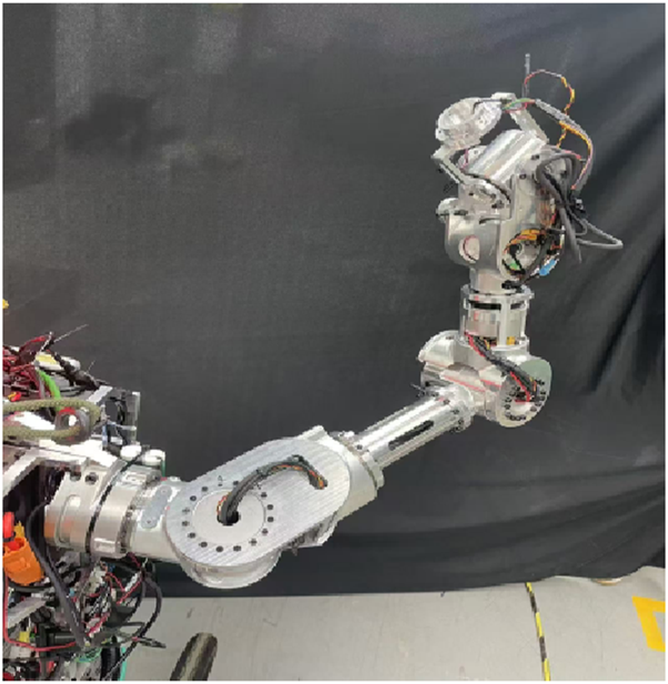 A robotic arm with exposed wiring and joints is positioned against a dark background. The arm features multiple articulated segments and visible cables, suggesting it is either under construction or undergoing repairs.