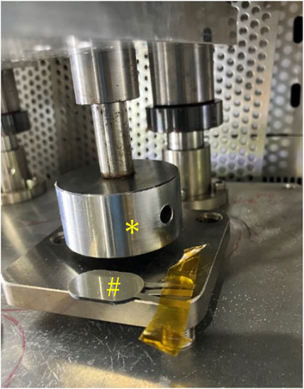 A metallic setup features a circular metal component marked with an asterisk and a smaller, disc-shaped structure marked with a hashtag attached to a flat rectangular base. The assembly is part of a larger machine setup, with a perforated metal background and yellow tape securing the disc to the base.