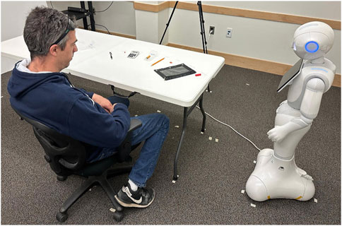 A man wearing a dark hoodie and jeans sits at a table facing a humanoid robot. The robot stands on a carpeted floor, with several objects like a calculator and pens on the table.