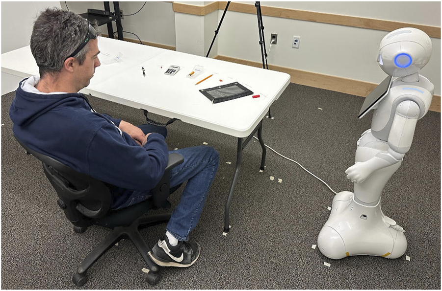 A man wearing a dark hoodie and jeans sits at a table facing a humanoid robot. The robot stands on a carpeted floor, with several objects like a calculator and pens on the table.