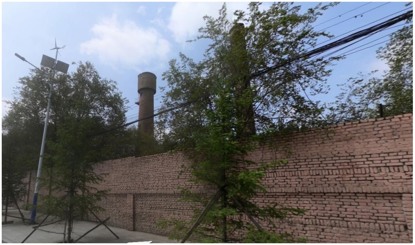 A brick wall with green trees and a utility pole topped with a wind turbine. Behind the wall, a tall cylindrical chimney is visible against a partly cloudy sky.