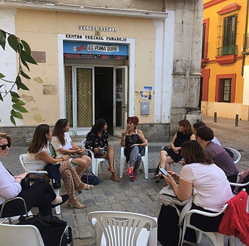 A group of people sitting in white plastic chairs arranged in a circle outside a building with the sign