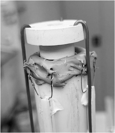 A close-up of a cylindrical object secured by metal hooks and wire. The top of the cylinder is covered with a layer of clay or putty-like material. The image is in black and white, focusing on the texture and composition of the materials.