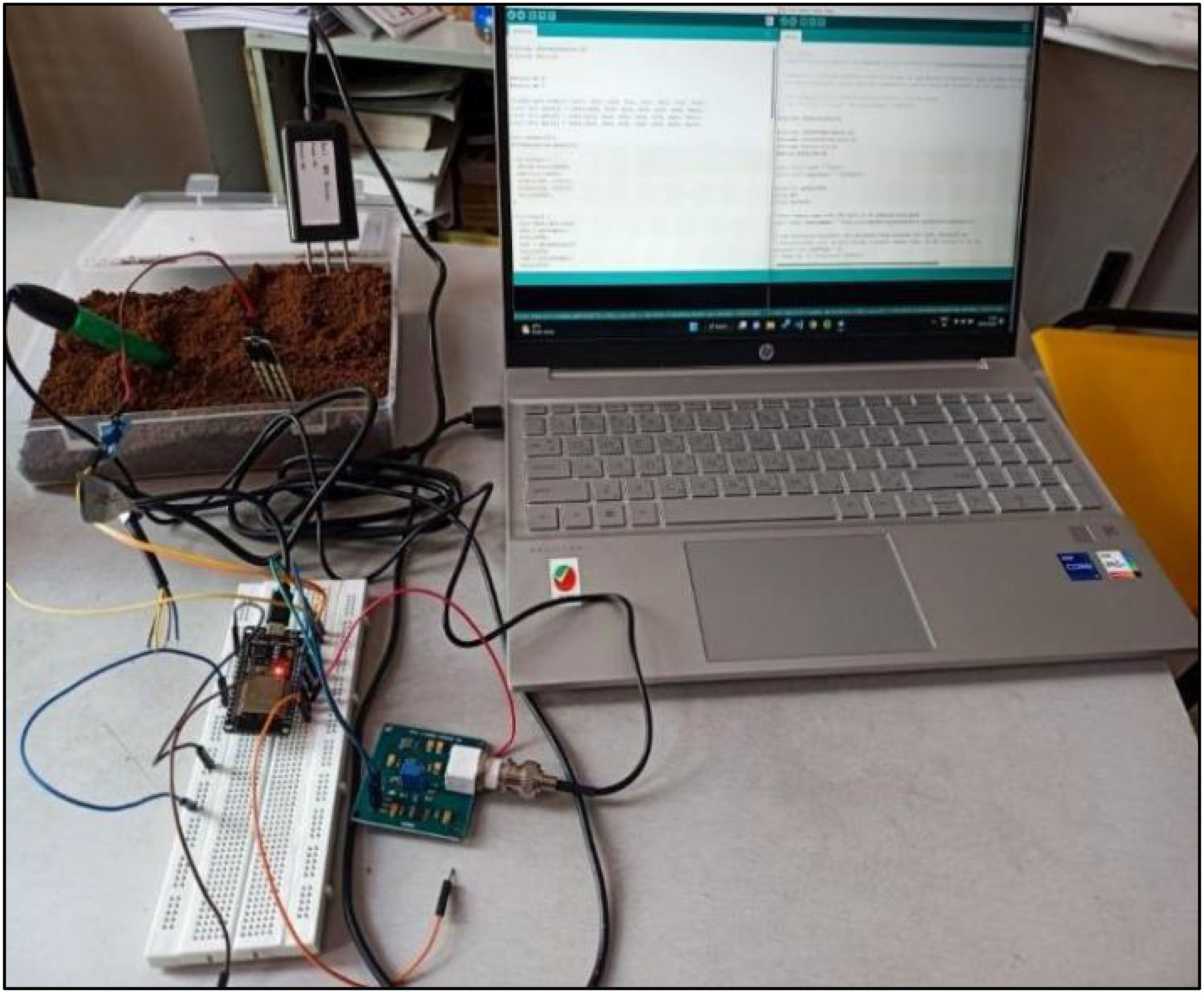 A laptop displaying code is connected to a soil moisture sensor setup. The setup includes a breadboard with wires and a microcontroller, linked to a container filled with soil where the sensor probe is inserted.