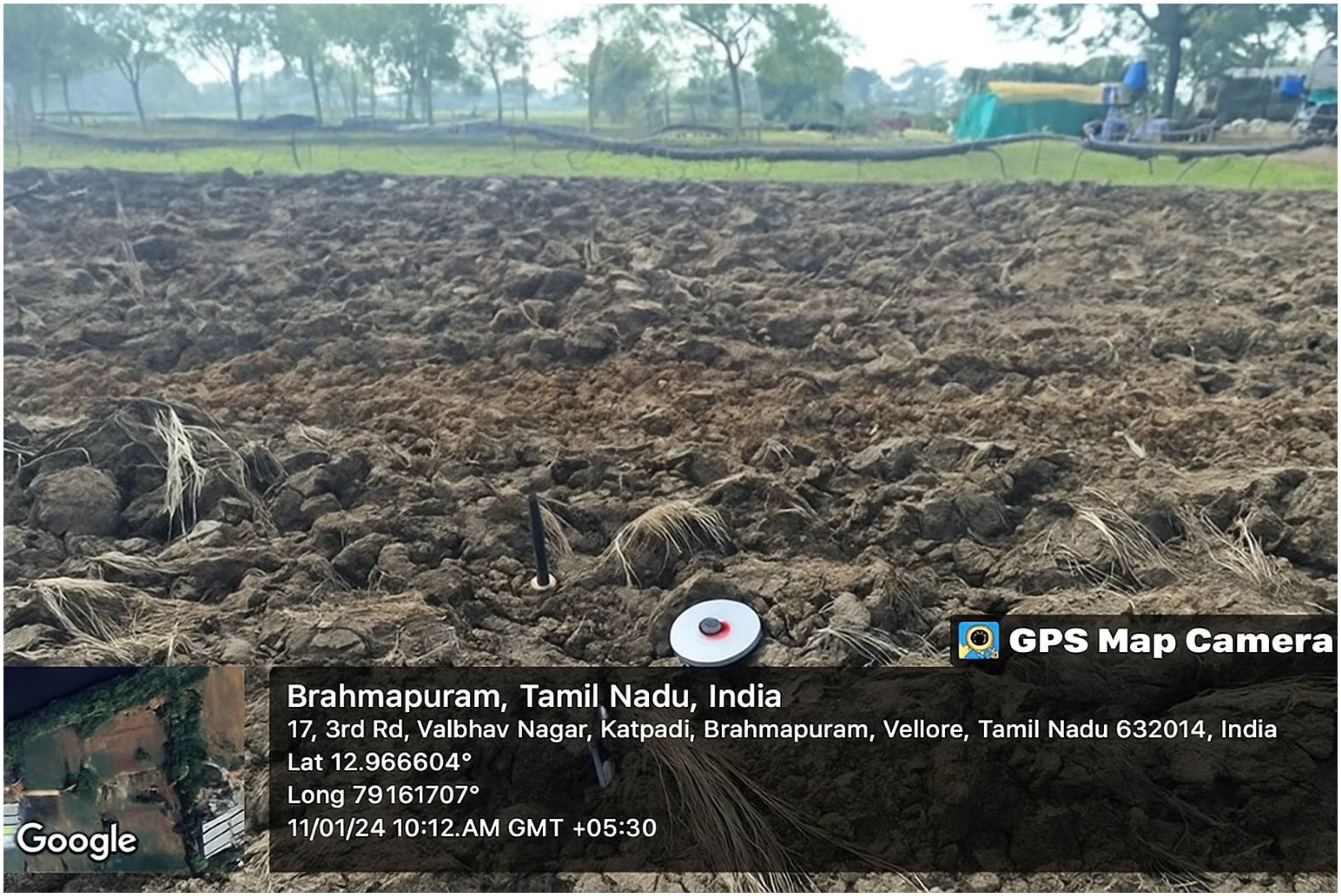 Field with freshly plowed soil, bordered by trees in the background. GPS Map Camera overlay shows location details: Brahmapuram, Tamil Nadu, India, with coordinates and timestamp as 11/01/24 at 10:12 AM GMT +5:30.