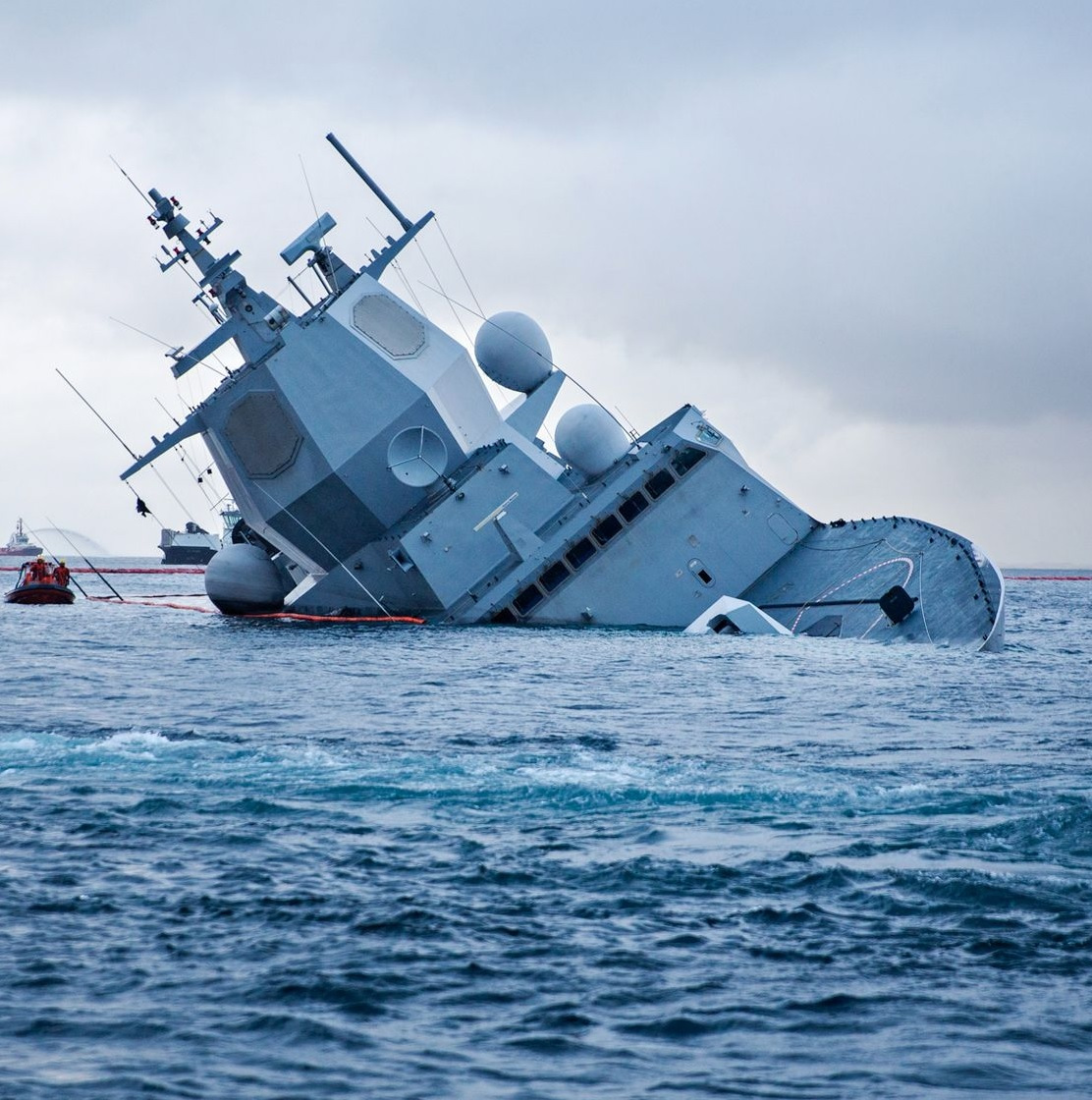 A large naval vessel partially submerged and tilting heavily to one side in a body of water. Nearby, rescue boats are present, suggesting a salvage or rescue operation. The sky is overcast, contributing to a somber atmosphere.