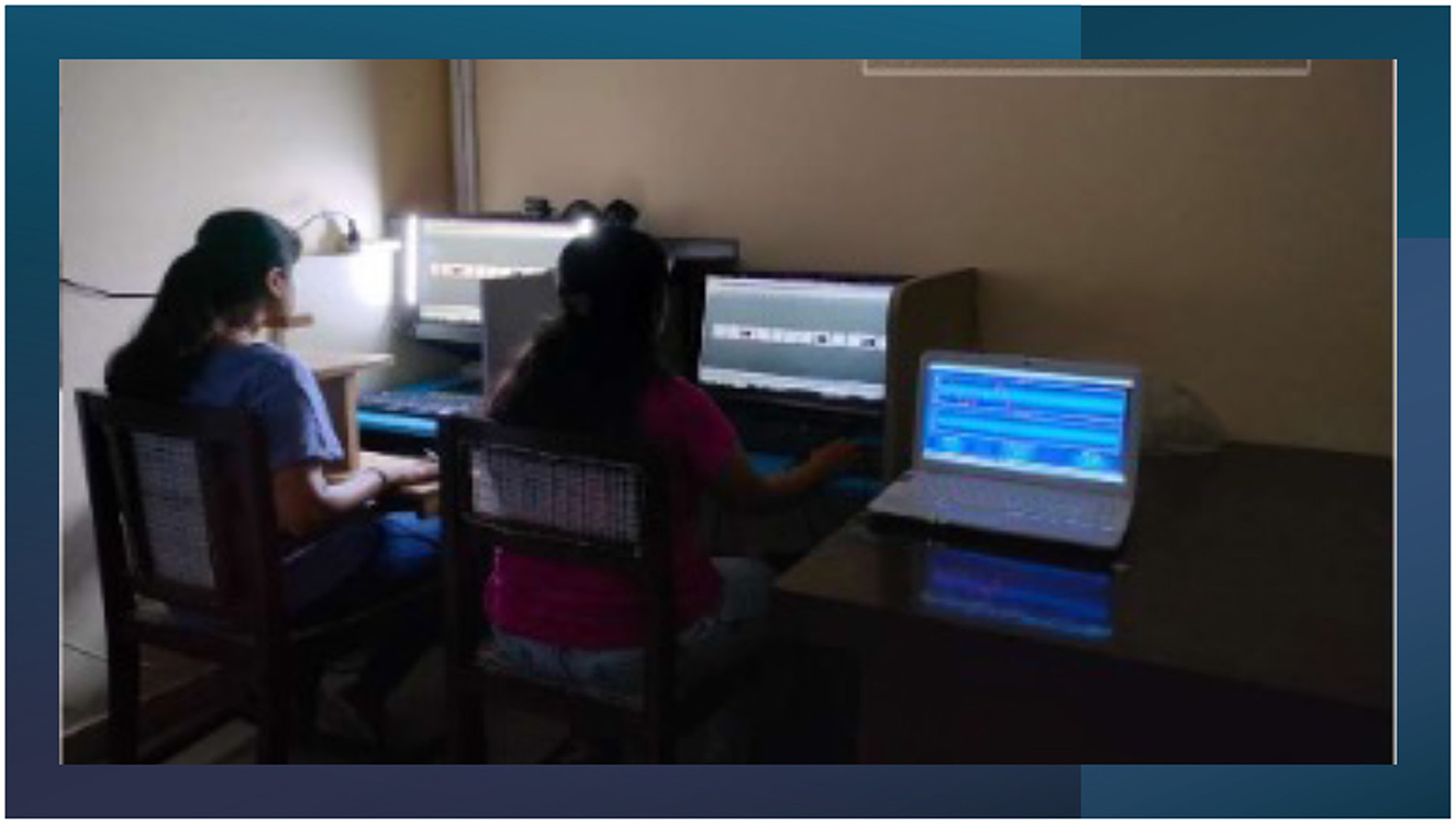 Two people sit at desks with computers displaying editing software. One laptop is on a separate table showing a blue screen with various data. The room is dimly lit.