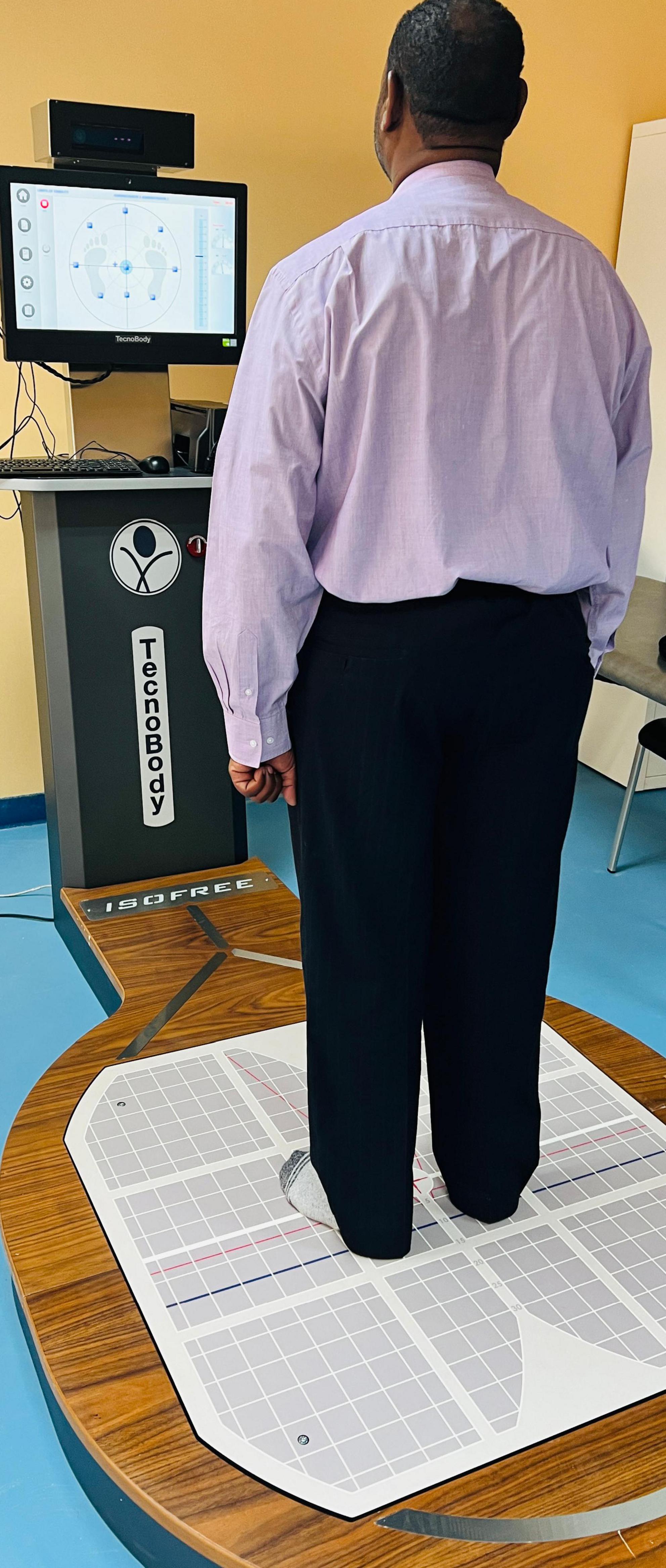 A person stands on a TecnoBody ISOBREE platform, facing a screen displaying a balance assessment interface. The platform features a detailed grid for precise positioning, and the setup suggests a balance or posture evaluation in a clinical setting.