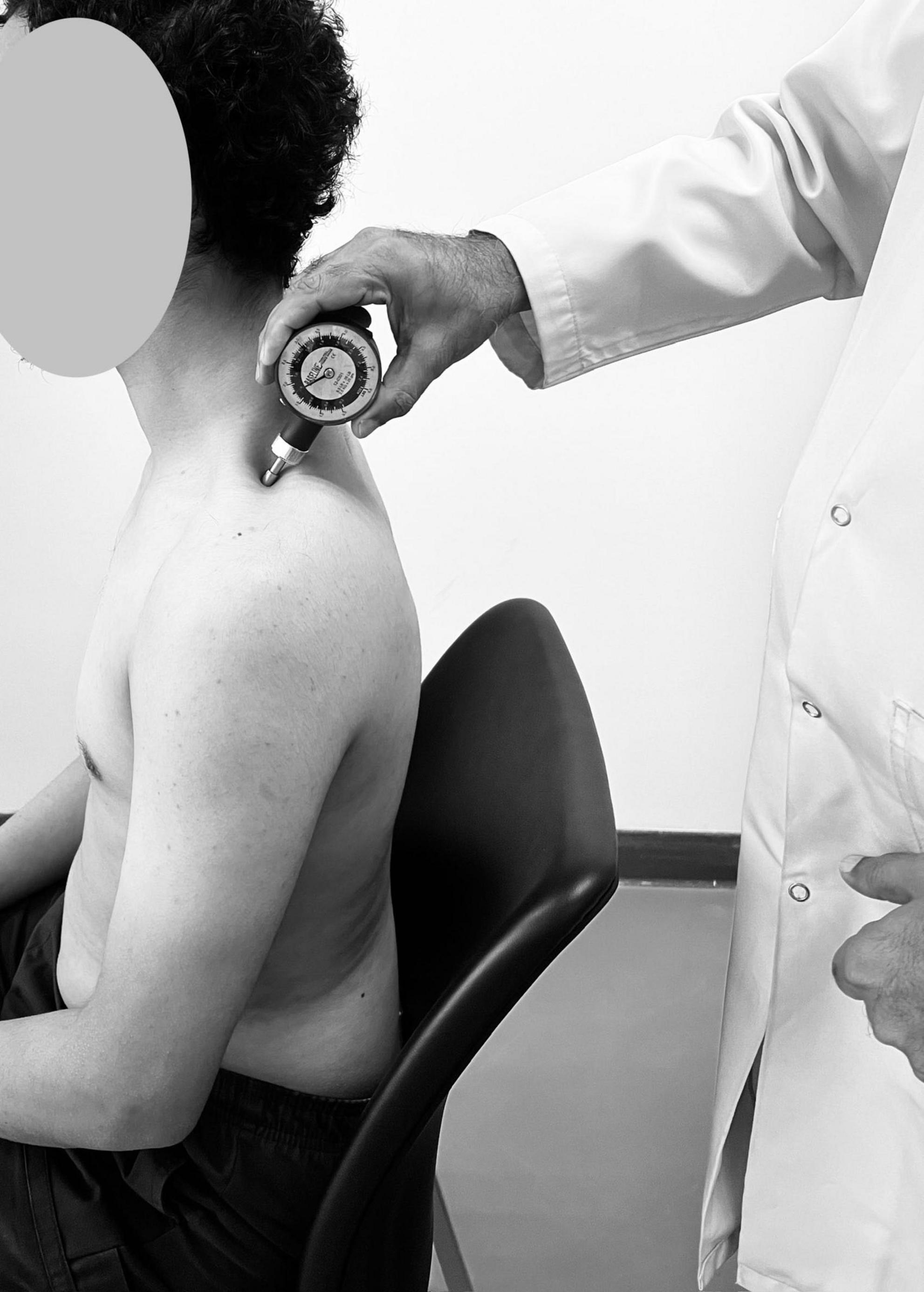 A healthcare professional in a lab coat uses a sphygmomanometer to measure the blood pressure of a seated male patient. The patient is shirtless, and the examination takes place in a clinical setting.