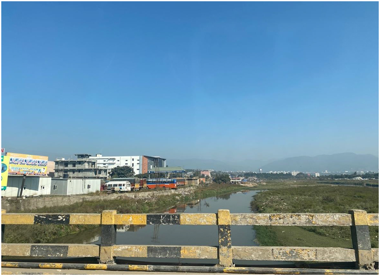 River flowing through a grassy area with buildings and industrial factories on the left, viewed from a bridge with yellow and black railings. A polluted river with brown, muddy waters flows below this bridge. Clear blue sky and distant hills in the background.