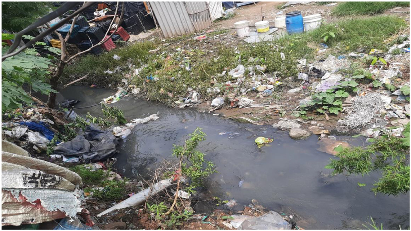 Polluted stream flowing through a cluttered area with scattered trash and debris. Vegetation grows along the banks, and various containers are visible in the background.