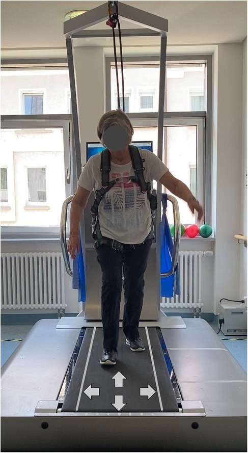 A person is walking on a treadmill equipped with safety harnesses and overhead support straps. The treadmill features directional arrows indicating movement directions. The setting appears to be a therapy or rehabilitation center with large windows and exercise equipment in the background.