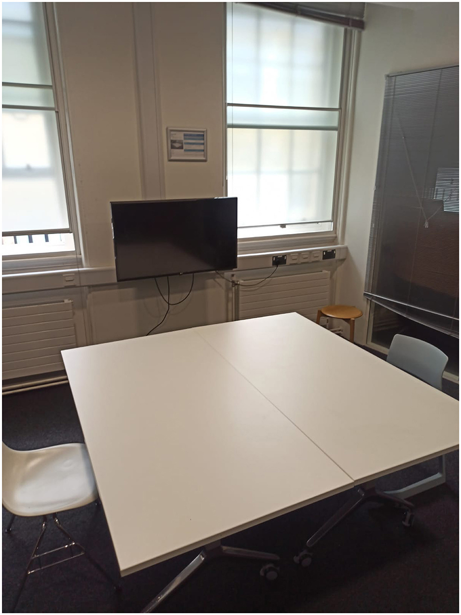 A small meeting room with a white rectangular table surrounded by two chairs and a stool. A wall-mounted television is connected to visible cables. Two windows with closed blinds allow some natural light into the room.