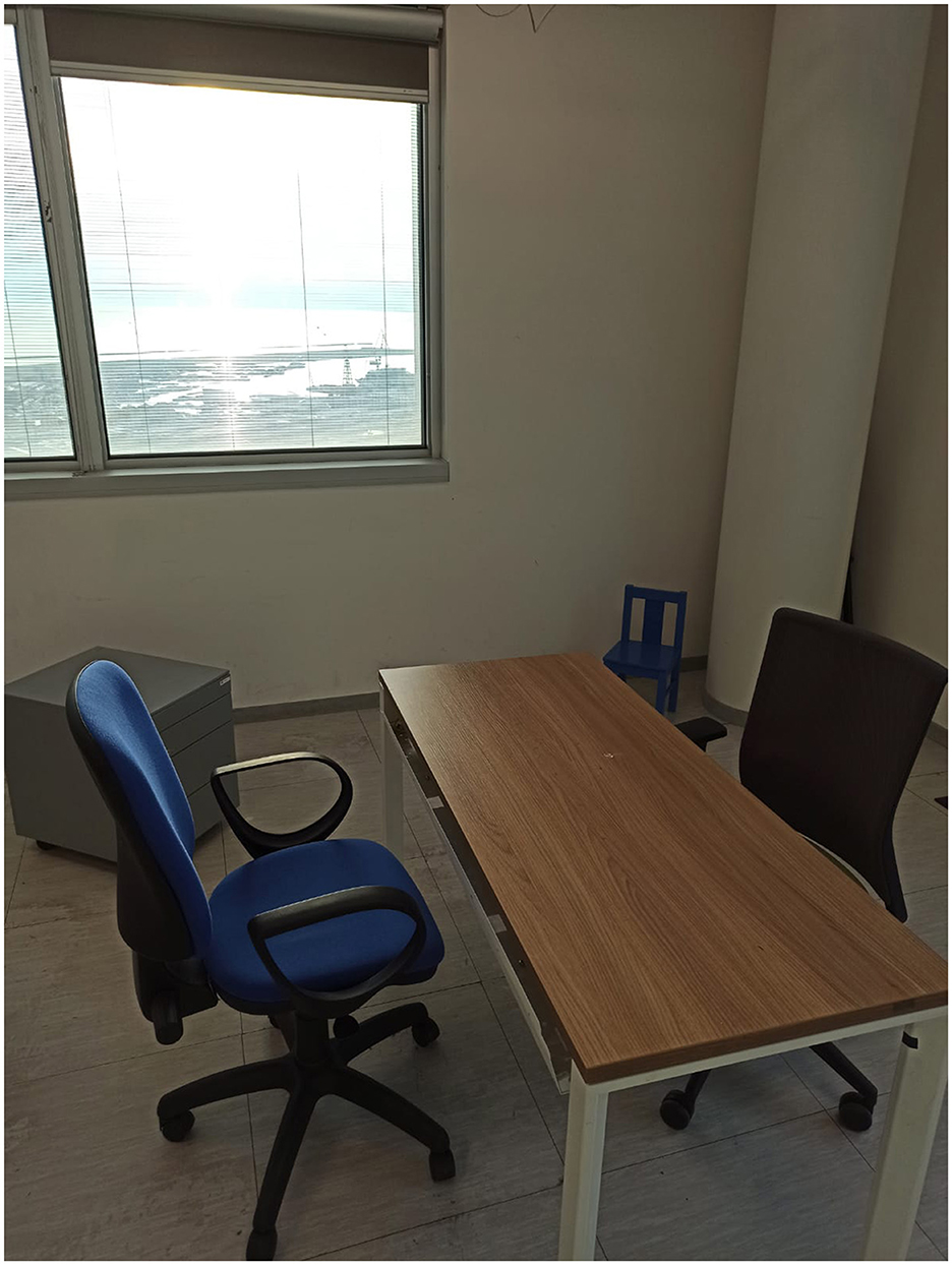 Office space with a wooden desk and two chairs, one with a blue cushion and the other black. A small blue chair is in the corner. A window reveals a bright outdoor view.