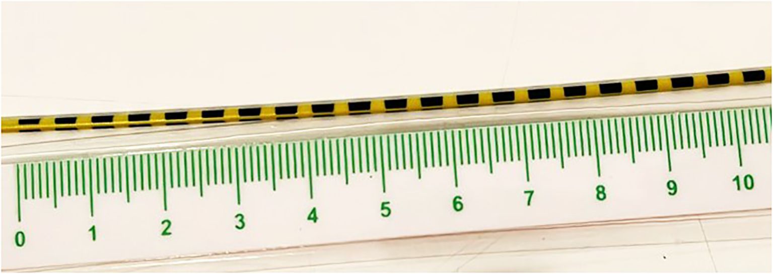 A thin black and yellow striped cylindrical object lies parallel to a transparent ruler with green markings. The ruler displays measurements from zero to ten, presumably in centimeters.