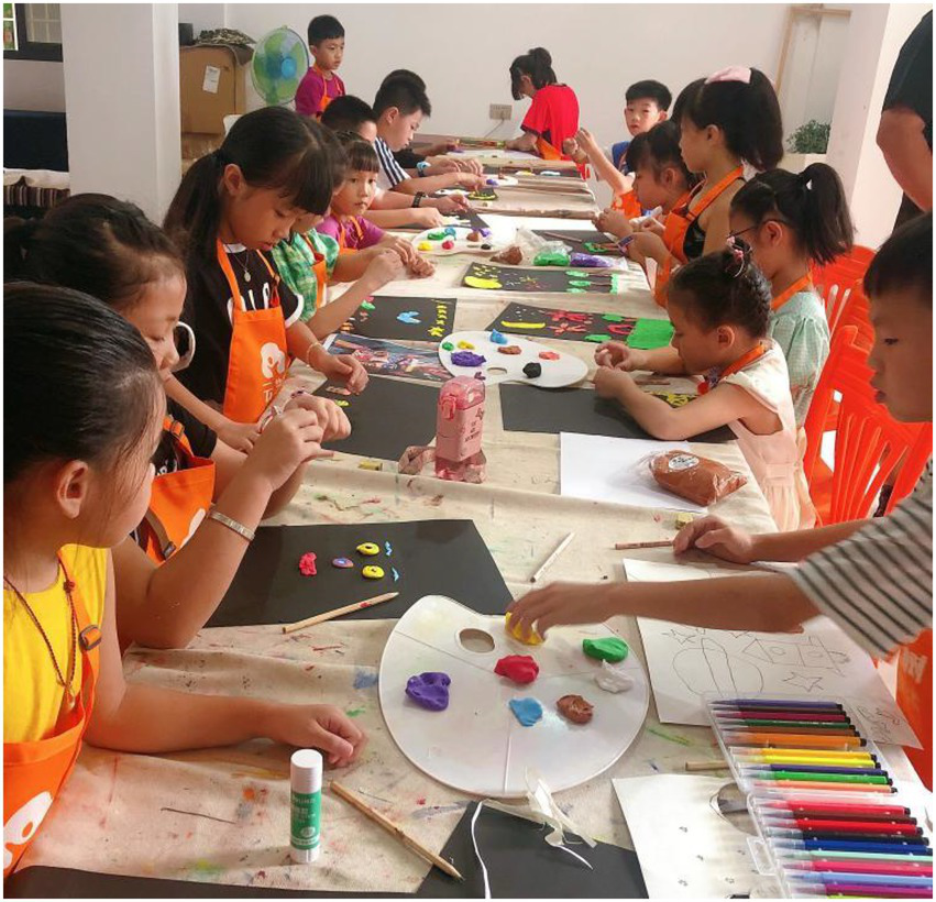 Children are seated at a table engaged in an arts and crafts session. They are using clay and colorful art supplies, creating designs on black paper. Bright orange chairs surround the table, and the atmosphere is lively and creative.