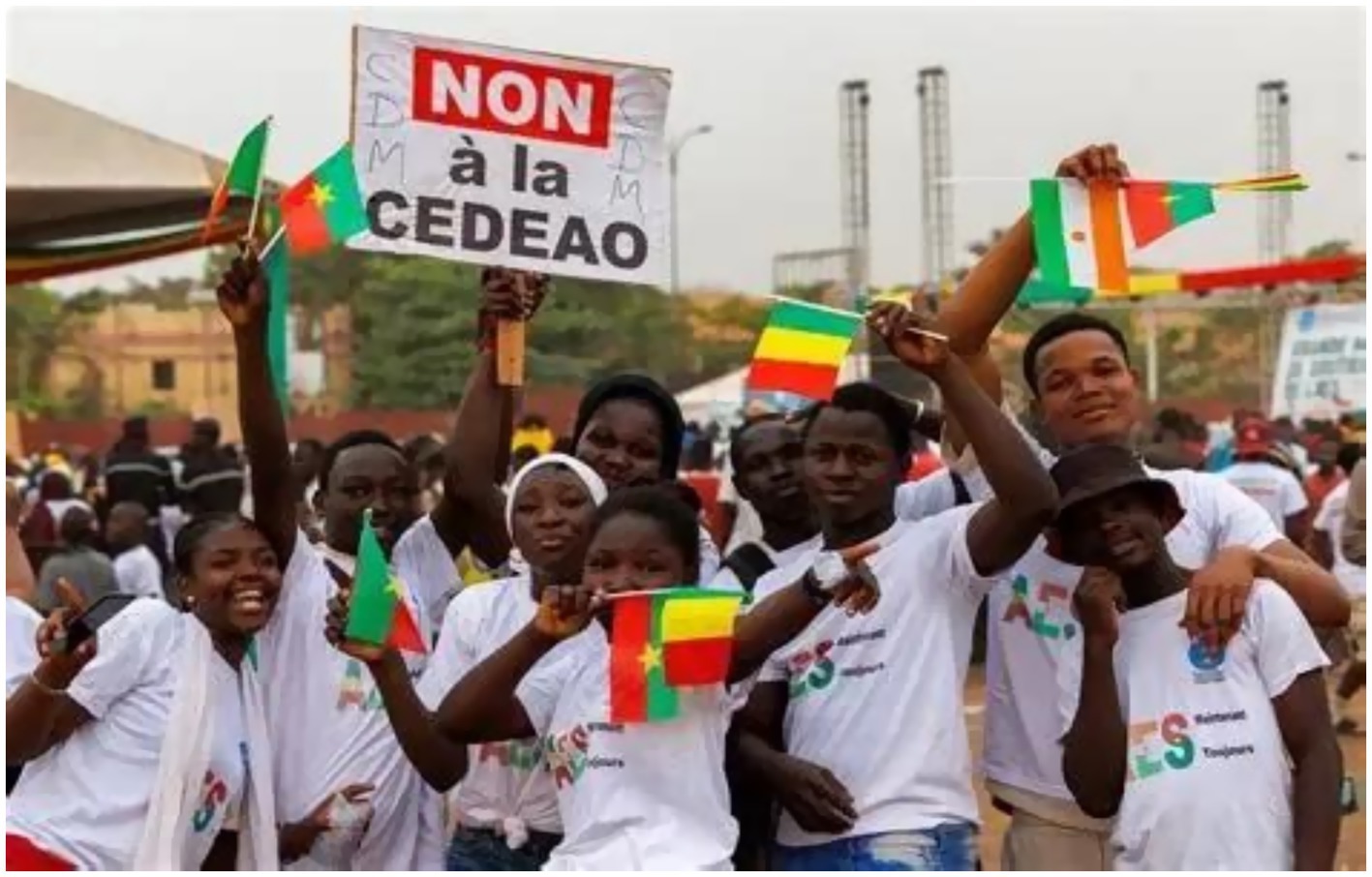 A group of people wearing white shirts gather at a demonstration, holding different national flags. One person holds a sign reading