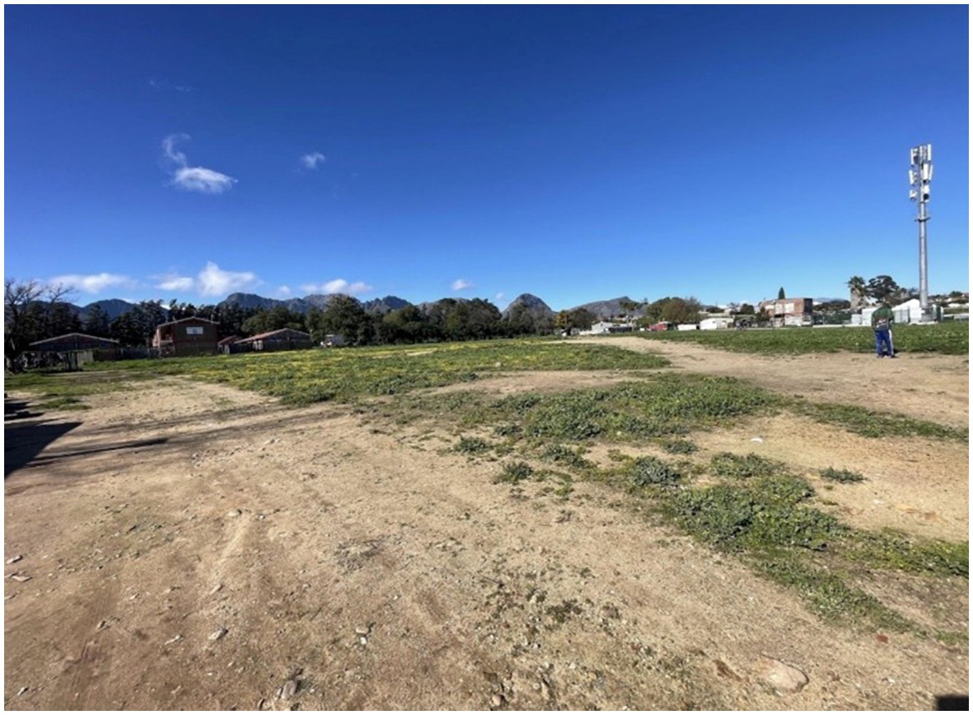 Open grassy field with patches of dirt under a clear blue sky. Buildings and trees are visible in the background, with mountains in the distance. A person stands near a tall pole on the right.