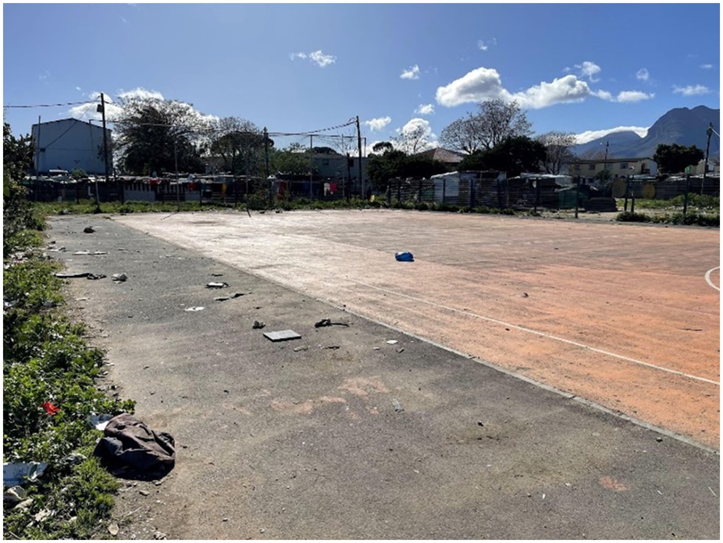 Outdoor basketball court with a worn surface, surrounded by overgrown grass and litter. In the background, shacks and a wire fence are visible, with mountains and a partly cloudy sky.