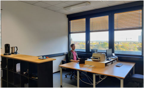A person working at a desk in an office with large windows. The desk has a laptop, monitors, and a printer. Nearby, there is a shelving unit with a coffee machine and kettle. The windows reveal a view of buildings and trees under a clear sky.