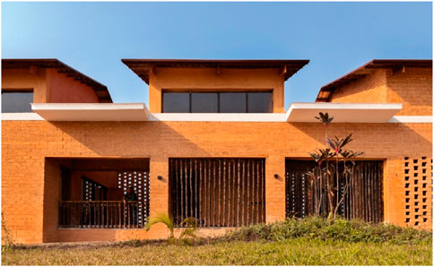 A modern house with an earthy orange facade and wooden slats across large windows. The structure features a flat roof and a small balcony with a railing. A plant is situated in front of the building on a grassy lawn.