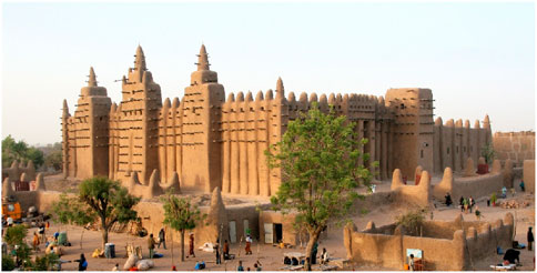 Large adobe building with tall, conical towers and wooden beams protruding from the walls, set in a sandy landscape with trees and people nearby.