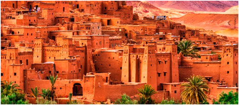 Ancient village of Ait Benhaddou, Morocco, showcasing traditional earthen clay architecture. The structures are stacked, with impressive towers, surrounded by palm trees against a backdrop of desert hills.