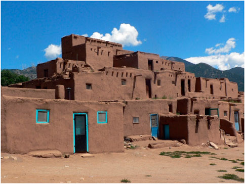 Adobe structures of a traditional pueblo village rise in tiers against a clear blue sky. The buildings are made of brown adobe, with blue-framed doors and windows, set against a mountainous backdrop.