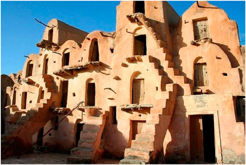 Traditional Berber granary with multiple arched, mud-brick structures featuring arched doorways and small windows. Steep external staircases lead to upper levels. The building is bathed in sunlight under a clear blue sky.
