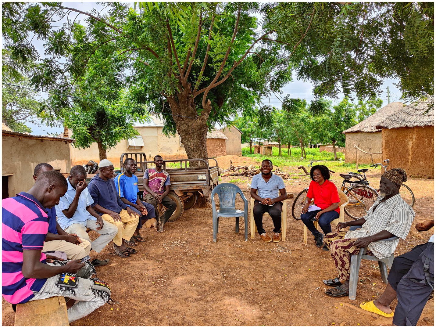 A group of people sit in a circle under a large tree in a rural setting. Some are on chairs and benches, while others stand nearby. Traditional huts and bicycles are visible in the background, and the ground is earthy. The atmosphere appears relaxed and communal.