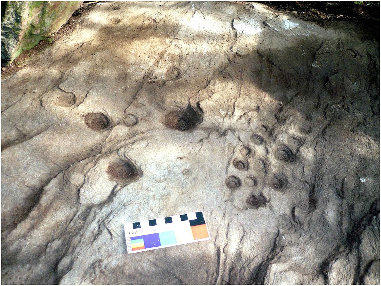 Rock surface featuring multiple small, circular depressions of varying sizes. A scale card with color blocks and measurements is placed near the depressions for reference. Shadows and textured rock are visible.