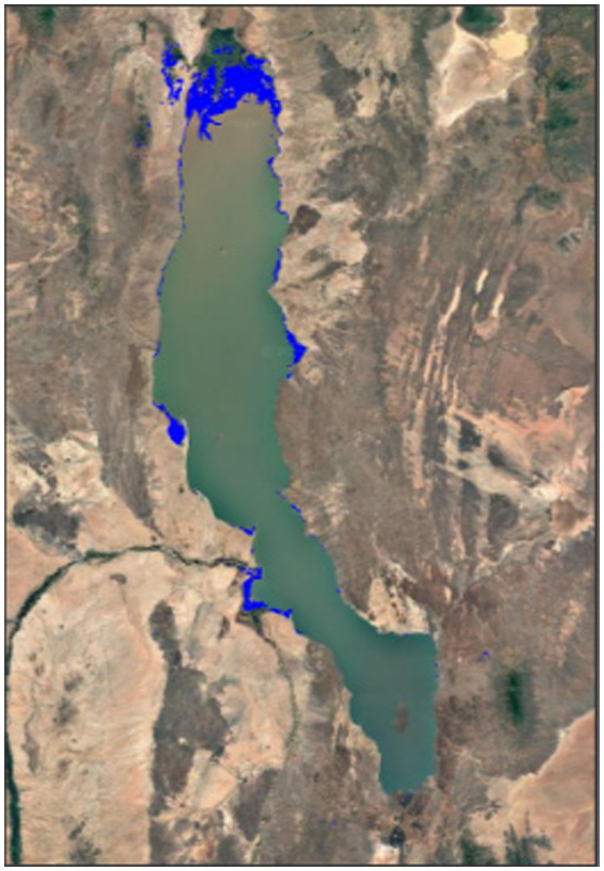 Satellite image showing a large, elongated lake surrounded by arid, rocky terrain. The lake appears greenish, with deep blue highlights along the edges. Surrounding areas are sandy and brown, with minimal vegetation.