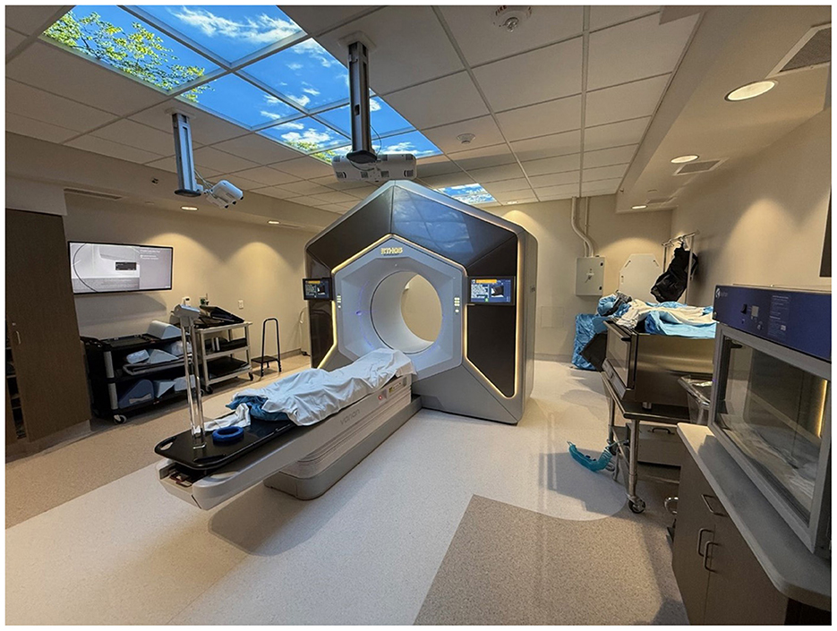 Medical imaging room with an advanced Ethos linac machine at the center. The machine has a circular opening and is surrounded by a control panel. A patient bed is positioned in front. The ceiling features panels with a sky image, and various medical equipment is present in the room.