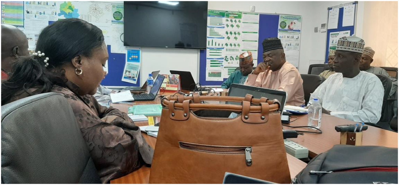 People are seated around a conference table, engaged in discussion. Laptops and documents are visible. The room has a wall with charts and a flat-screen TV. A brown handbag is placed prominently on the table.