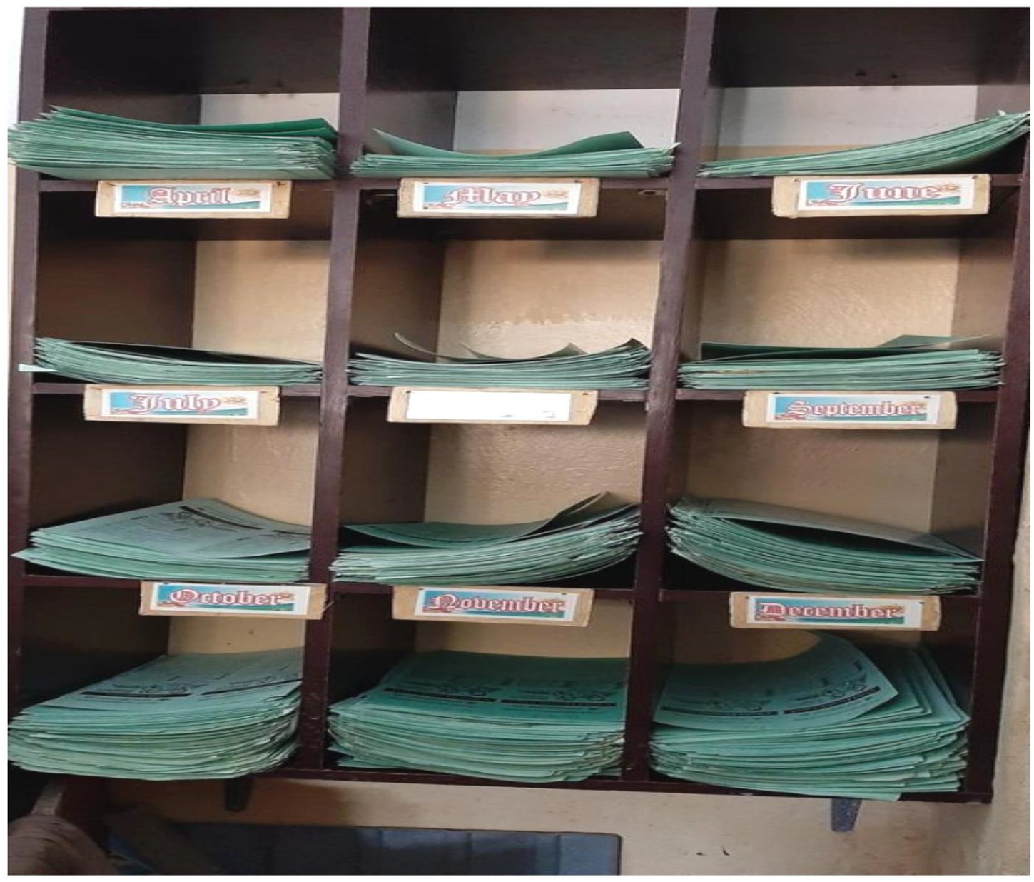 Wooden shelf with compartments labeled by months from April to December, each holding stacks of green paper sheets. April to June are on the top row, July to September in the middle, and October to December on the bottom.