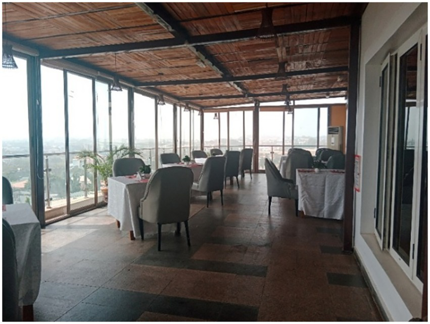 Sunny indoor restaurant with large windows offering a panoramic view. Tables are covered with white tablecloths, surrounded by gray chairs. Wooden ceiling and tiled floor create a warm atmosphere.