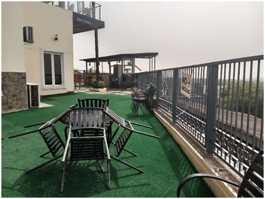 Outdoor terrace with artificial grass flooring, featuring a stack of upside-down metal chairs and tables. A building with large windows is on the left. An adjacent railing has a "No Smoking" sign. In the background, there is a shaded seating area.