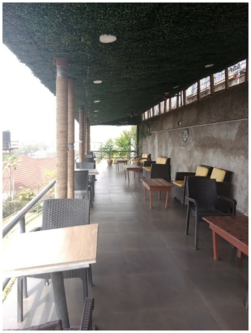 Outdoor terrace with wicker chairs and tables lining one side. A green artificial hedge covers the ceiling. Potted plants and a dart board adorn the space, overlooking a view of rooftops.
