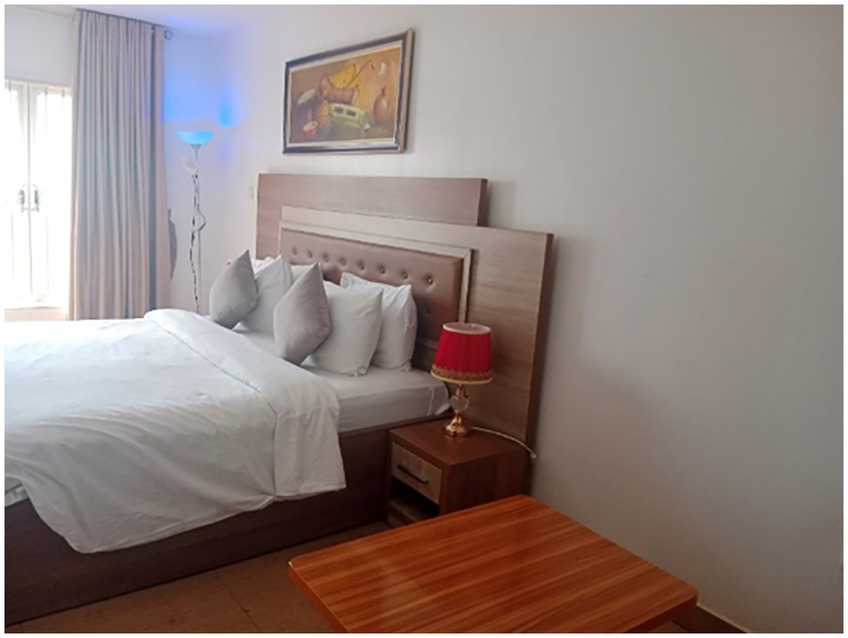 A well-lit bedroom features a neatly made bed with white linens and several gray pillows. A nightstand with a red lamp is beside the bed. A floor lamp and framed artwork are visible. Beige curtains cover a window, and a wooden table is in the foreground.