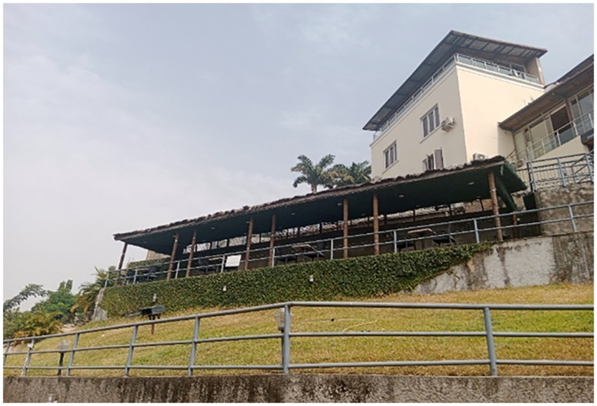 A multistory building with a slanted roof and a balcony sits on a grassy hill. A wooden structure with a thatched roof is partially visible in front of the building. Metal railings line the slope, and palm trees are seen in the background under a cloudy sky.
