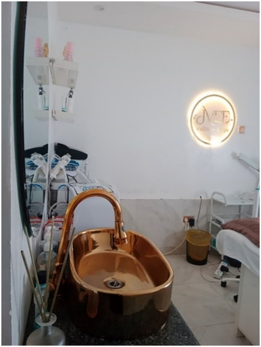 A modern spa room featuring a shiny bronze sink with a tall faucet on a dark countertop. Wall shelves with bottles are visible above the sink. A spa bed and equipment are in the background. A circular illuminated sign with text is on the wall.