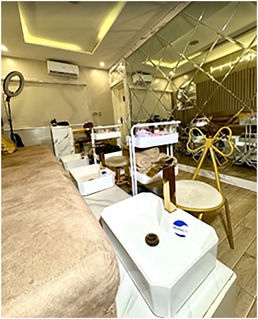 Modern salon interior with mirrored walls, warm lighting, and plush seating. White pedicure stations are arranged neatly with towels and beauty products. A ring light and decorative chair accent the space.