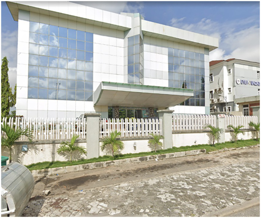 Modern glass-fronted building with a white facade and large windows, reflecting clouds. It has a protruding entrance and is surrounded by a white fence with small palm trees along the perimeter.
