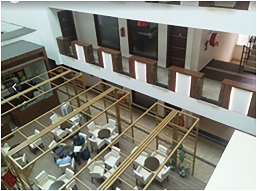 Atrium view of a multi-story building featuring a dining area with white tables and chairs arranged below a partially open lattice structure. Upper floors have wooden doors and paneling.