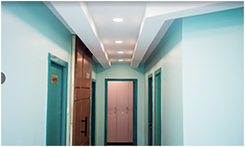 A narrow hallway painted in light teal with a white ceiling featuring recessed lighting. There are doors along the sides, and a closed wooden door at the end of the corridor.